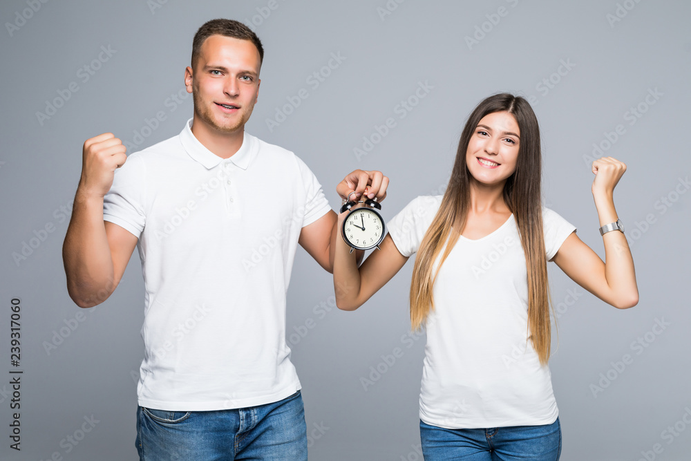 Portrait of happy young loving couple holding alarm clock make winner gesture isolated on gray background Portrait of happy young loving couple holding alarm clock make winner gesture isolated on gray background