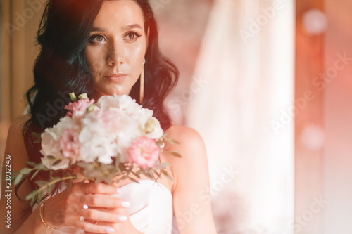 Pretty young caucasian bride with bouquet of flowers posing indoor. Lovely girl with spanish appearance closeup toned  home portrait. Dark eyed bride in white dress with pensive face tender fellings.