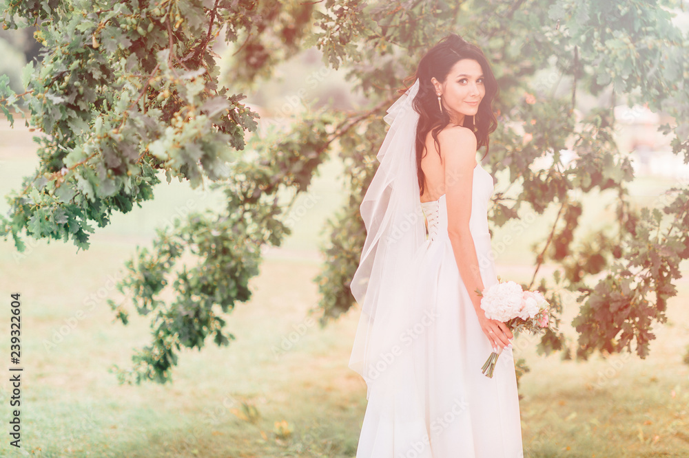 Happy tanned bride in beautiful white dress posing with boquet of flowers on green abstract background in summer day. Bridal outdoor photosession. Beautiful cheerful brunette bride lifestyle portrait. Happy tanned bride in beautiful white dress posing with boquet of flowers on green abstract background in summer day. Bridal outdoor photosession. Beautiful cheerful brunette bride lifestyle portrait.
