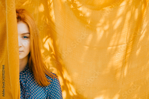 фотография Beautiful girl with red hair hiding half face behind orange blanket with abstract shadows on background