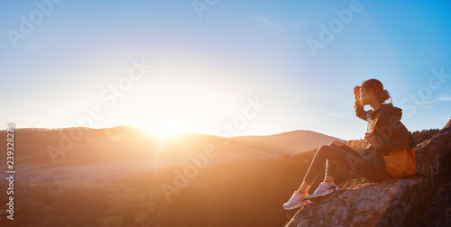 woman sits on edge of cliff against background of sunrise