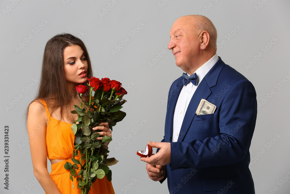 Senior rich man proposing to young woman on grey background. Marriage of convenience Senior rich man proposing to young woman on grey background. Marriage of convenience