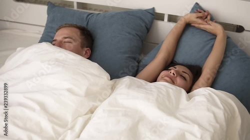 Young smiling brunette woman waking up happy after healthy sleep stretching on comfortable bed with sleeping man near, refreshed girl enjoys pleasant wakeup, good morning in cozy loft bedroom and