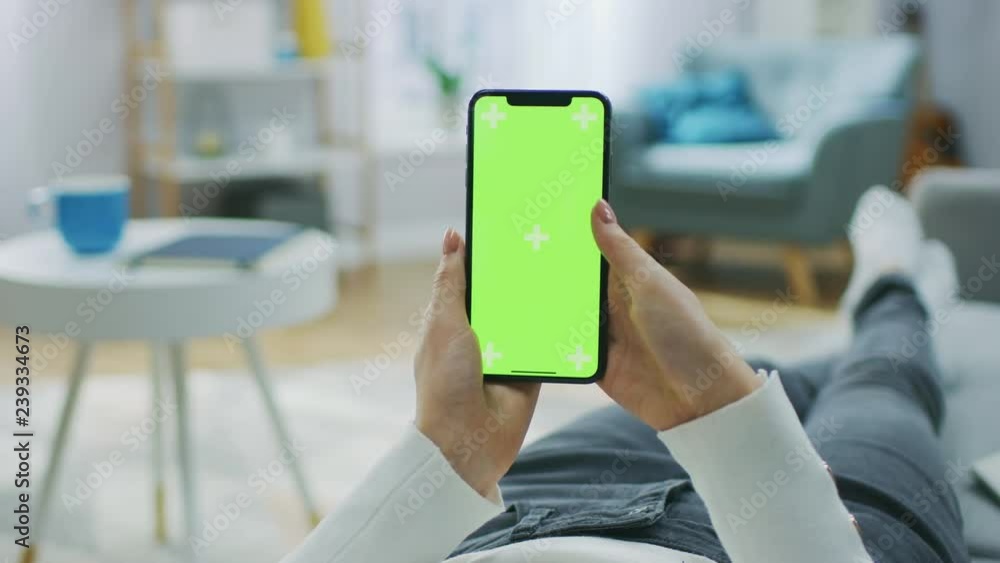Woman at Home Lying on a Couch using Smartphone with Green Mock-up Screen, Doing Swiping, Scrolling Gestures. Girl Using Mobile Phone, Internet Social Networks Browsing. Point of View Camera Shot. Woman at Home Lying on a Couch using Smartphone with Green Mock-up Screen, Doing Swiping, Scrolling Gestures. Girl Using Mobile Phone, Internet Social Networks Browsing. Point of View Camera Shot.