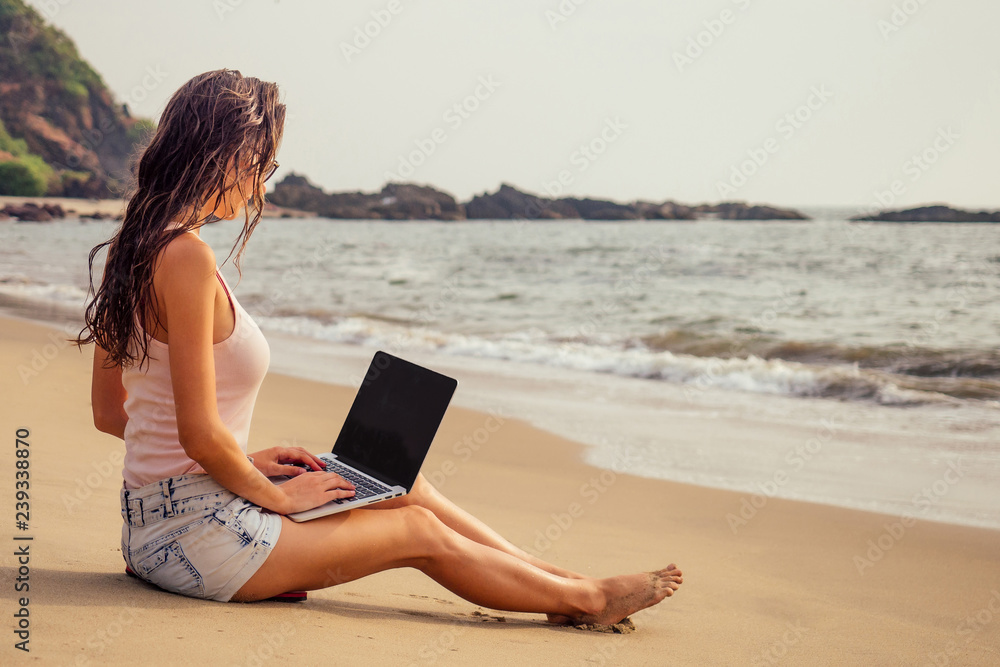 young woman with laptop on tropical vacation sitting on the sand on a desert paradise island on the beach by the sea.girl freelancer surfer on the Indian Ocean.remote work and freedom freelancing.