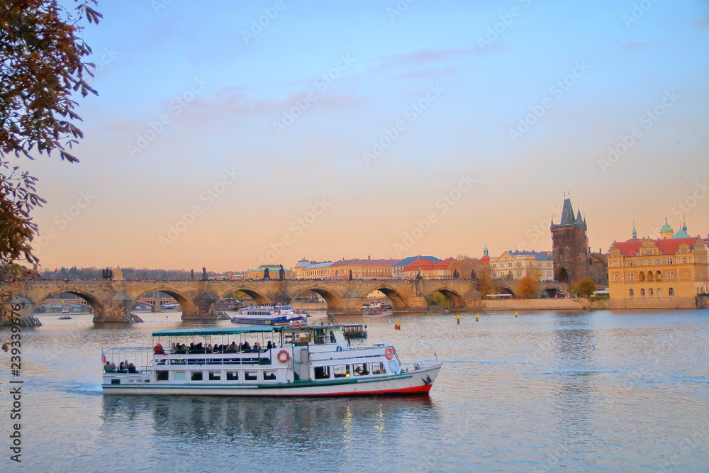 Naklejka premium Pleasure boat on the Vltava River in Prague against the backdrop of Charles Bridge at sunset.
