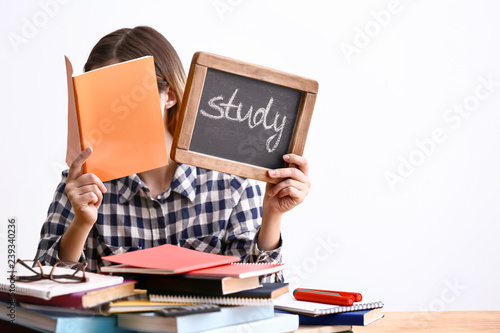 Young woman sitting at table with a lot of books and holding chalkboard with word STUDY against white background