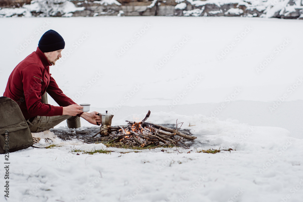 Obraz premium Traveling man a sits near camp fire an in winter time and pours itself hot coffee . Concept adventure active vacations outdoor. Winter camping