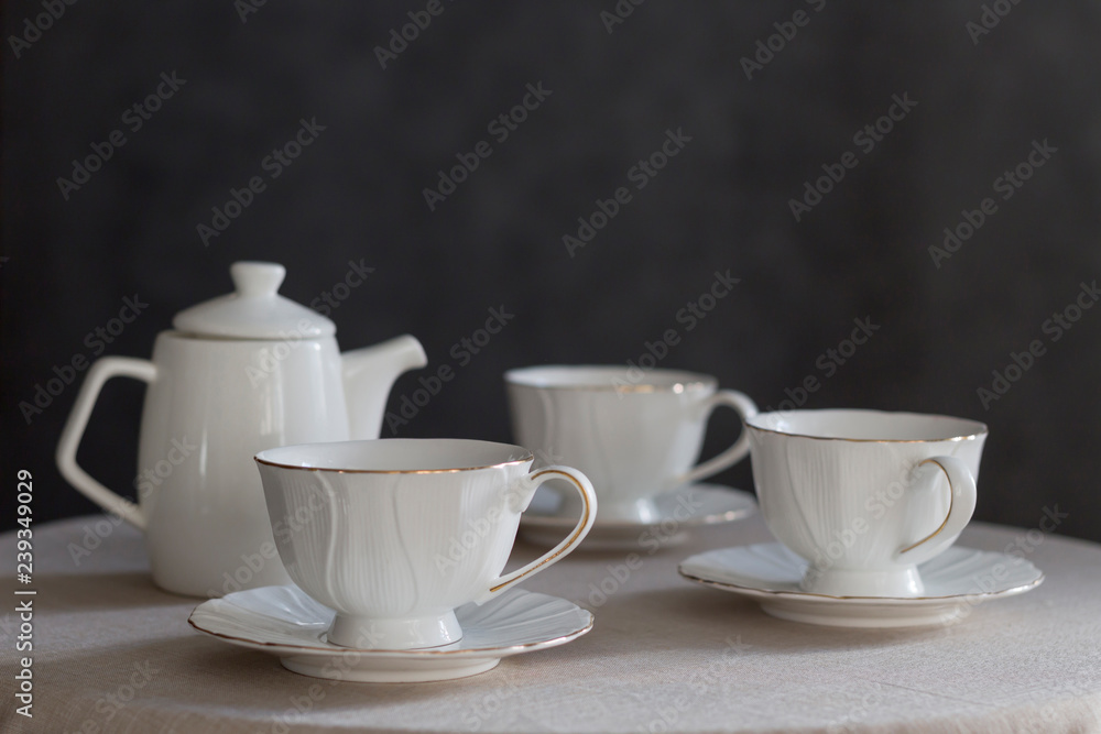 Tableware for tea party - teapot and cups - on white tablecloth on dark gray background. Close-up, copy space Tableware for tea party - teapot and cups - on white tablecloth on dark gray background. Close-up, copy space