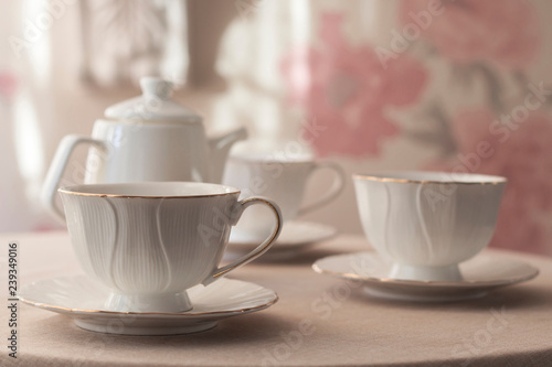 Tableware for tea party - teapot and cups - on white tablecloth on light pink floral background with sunlight. Close-up, copy space