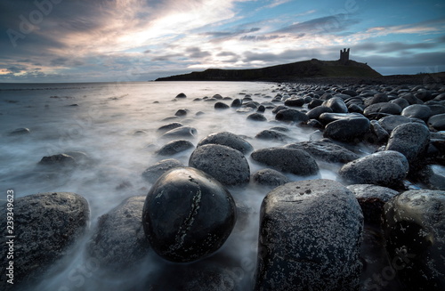 View at sunrise towards the ruin of Dunstanburgh Castle from the beach of basalt boulders known as The Rumble Churn at the southern end of Embleton Bay, Embleton, near Alnwick, Northumberland, England, United Kingdom, Europe