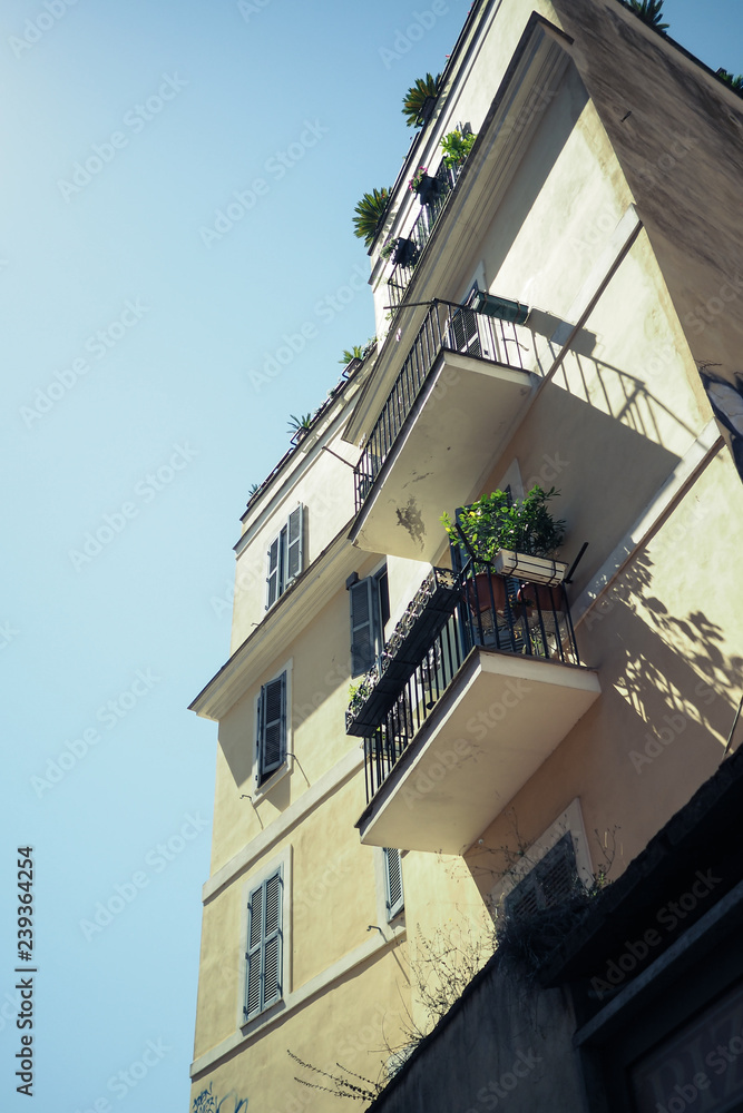 Pale yellow building with metal juliet balconies and garden boxes against a blue sky, in Rome, Italy