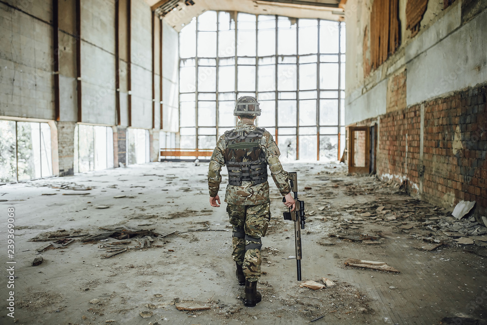 Military reconnaissance operation! A young soldier with a big rifle in his hands goes through a collapsed building, a view from the back! Military reconnaissance operation! A young soldier with a big rifle in his hands goes through a collapsed building, a view from the back!