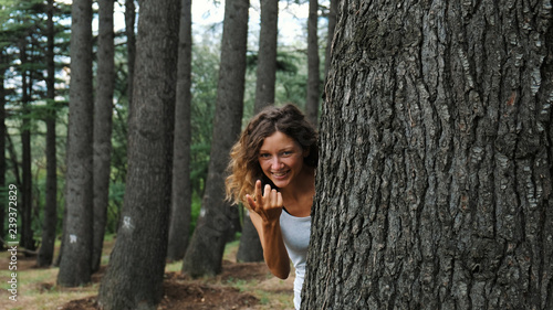 A girl looks out from behind a tree and beckons a finger to her.