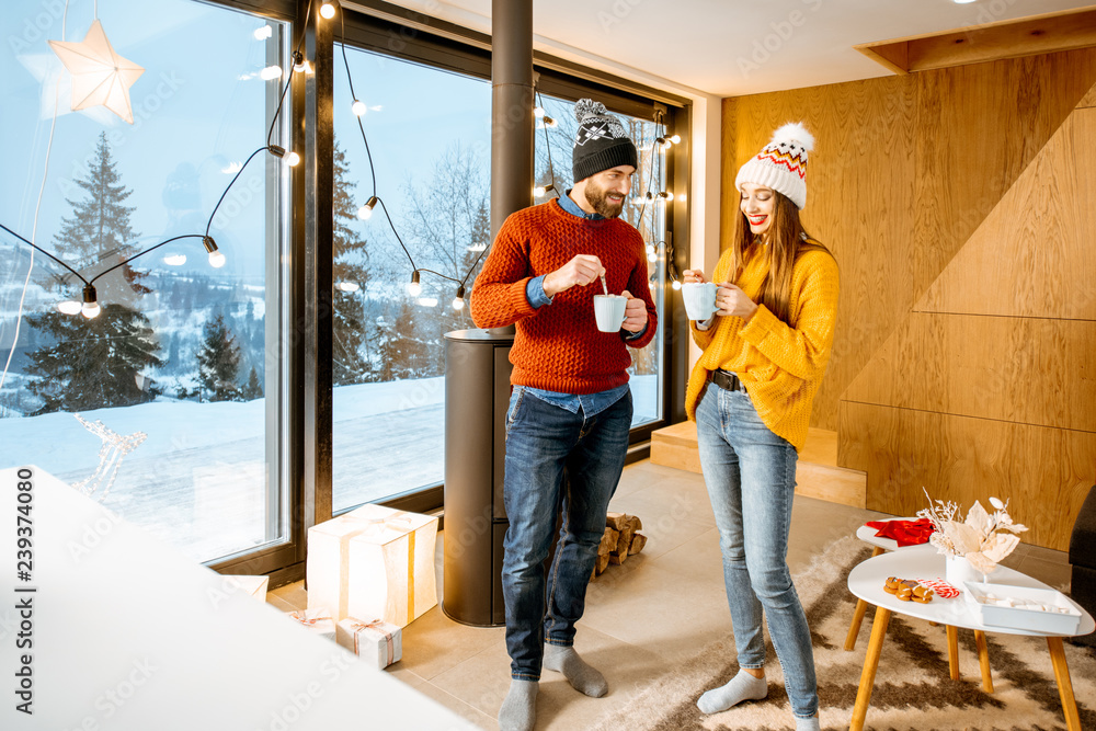 Young couple dressed in bright sweaters and hats standing together with hot drinks near the fireplace in the modern house durnig winter time Young couple dressed in bright sweaters and hats standing together with hot drinks near the fireplace in the modern house durnig winter time