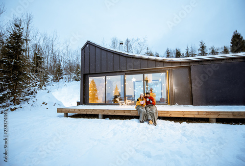 Young couple warming up with plaid and hot drinks sitting on the terrace of the modern house in the mountains durnig the winter holidays. Wide landsacpe view