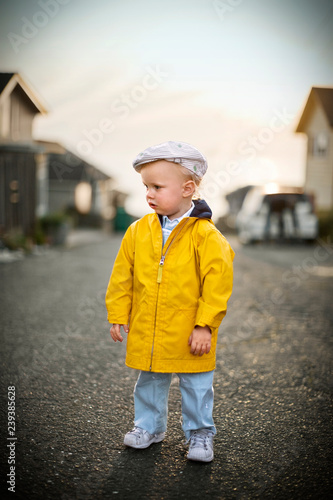 Young toddler wearing a flat cap and yellow raincoat while standing on a suburban street. photo