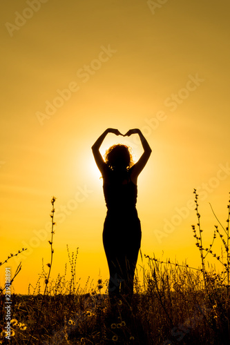 A young woman dancing at sunset in a field among the grass and holding her hands over her head in the shape of a heart. Silhouette. Vertical orientation of the image