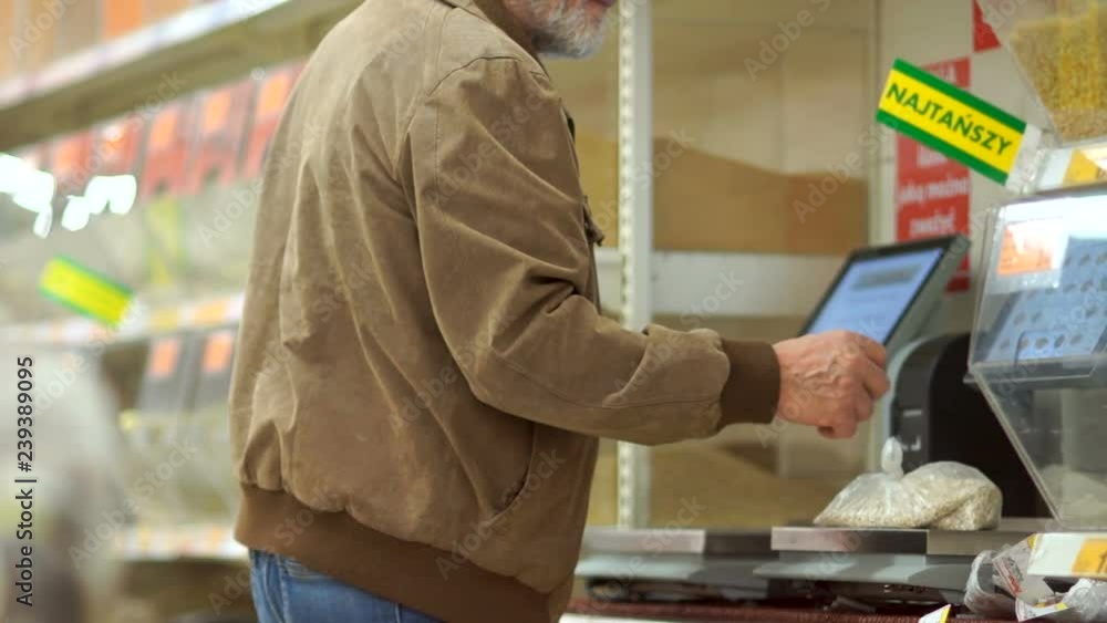 Gray-haired retiree with a beard weighs cereals on scales in a supermarket, sticks a price tag on a package. Lonely old man, grocery department Gray-haired retiree with a beard weighs cereals on scales in a supermarket, sticks a price tag on a package. Lonely old man, grocery department