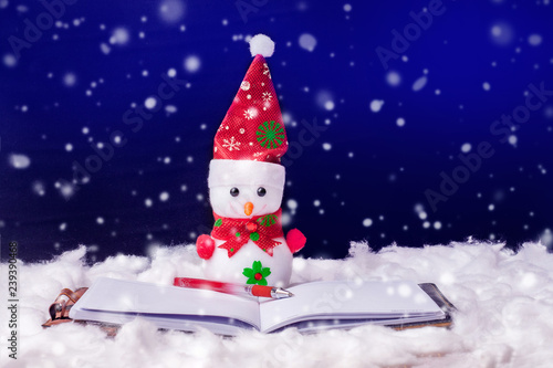 Toy snowman in front of an open book. Fiction reading. Snowman on a dark background during the snowfall. Happy Christmas and New Year s greetings_