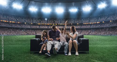 A family is watching a soccer moment, sitting on the couch located in the middle of the soccer stadium.