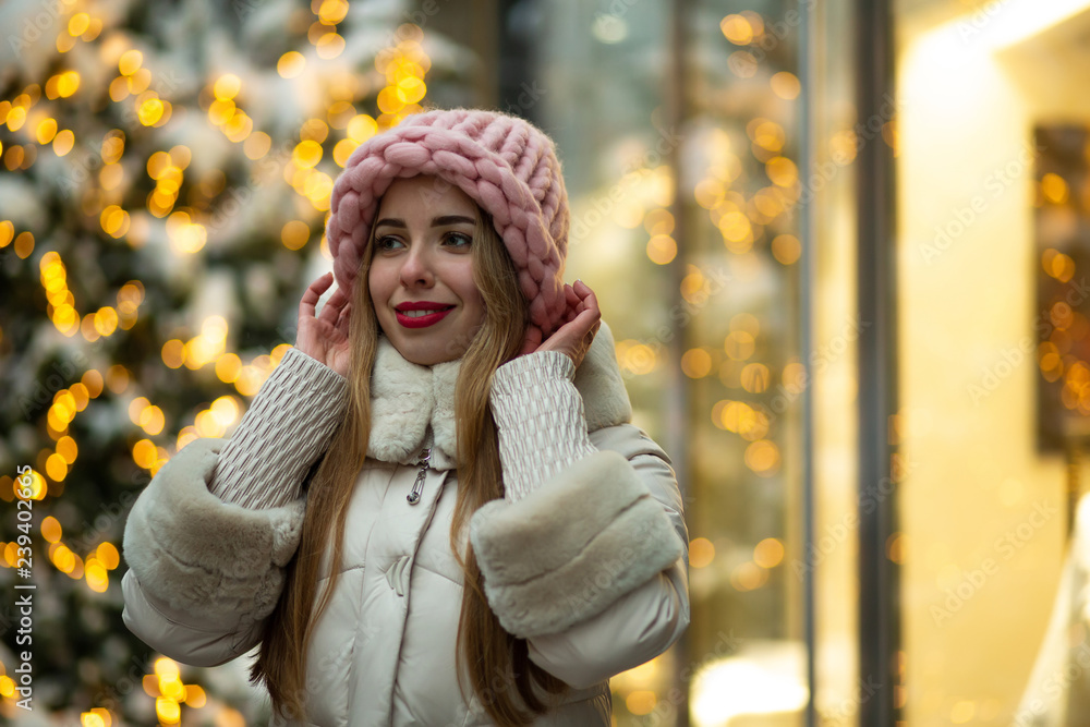 Glorious blonde model with red lips wearing volumetric hat and winter coat walking down the street decorated with garlands. Space for text Glorious blonde model with red lips wearing volumetric hat and winter coat walking down the street decorated with garlands. Space for text