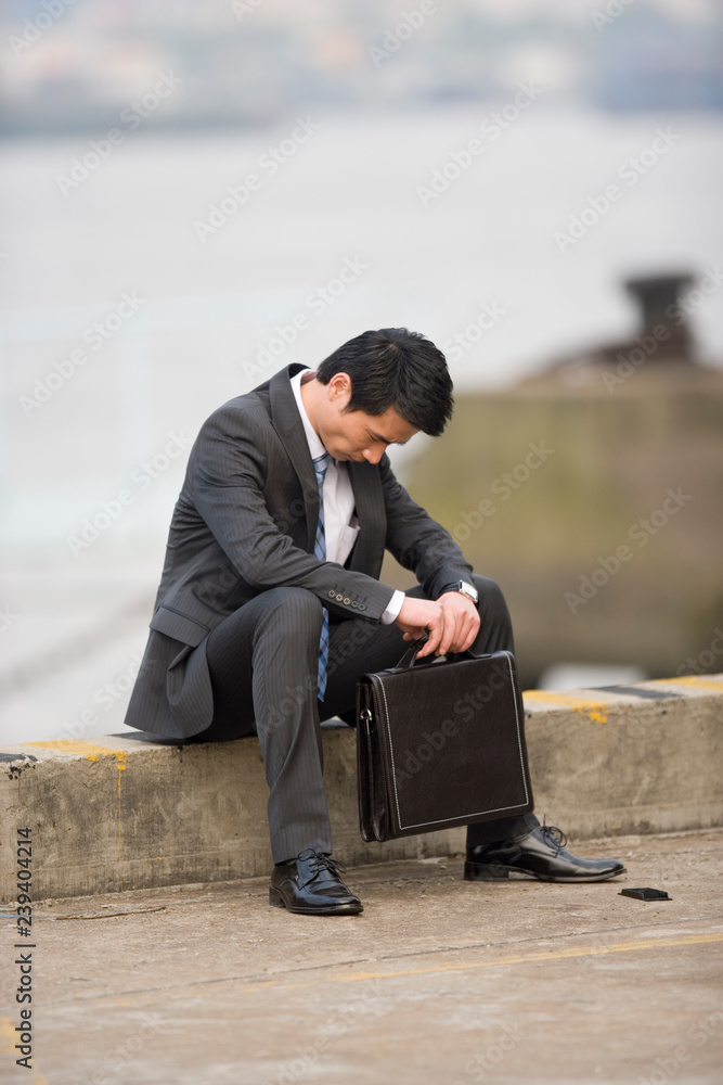 Young adult businessman sitting on the end of a wharf with his head bowed.