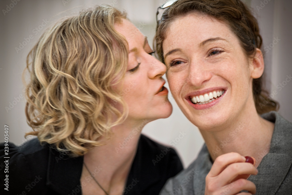 © Erickson Stock - Smiling young woman being kissed by her girlfriend on the cheek. © Erickson Stock - Smiling young woman being kissed by her girlfriend on the cheek.