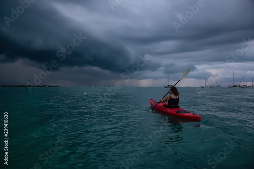 Adventurous girl on a red kayak is kayaking towards a thunderstorm during a dramatic sunset. Taken in Key West, Florida Keys, United States.