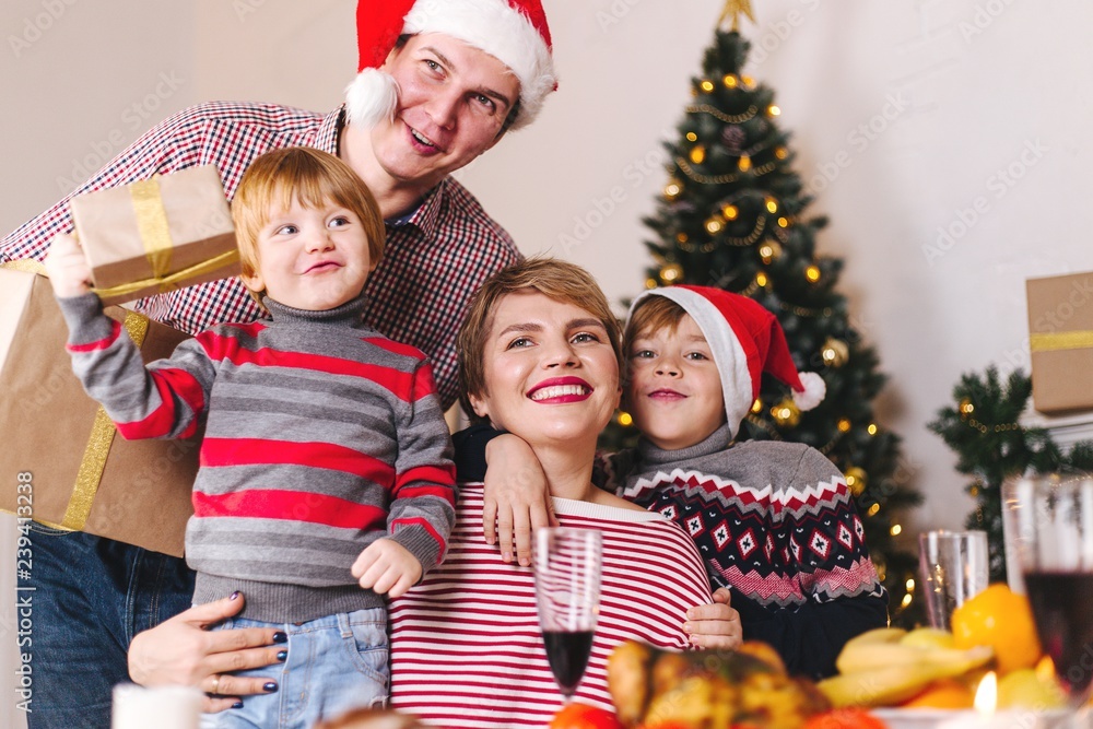 Happy family parents with children in Santa Claus hats stand at the Christmas table with gifts. Joyful family holiday Happy family parents with children in Santa Claus hats stand at the Christmas table with gifts. Joyful family holiday