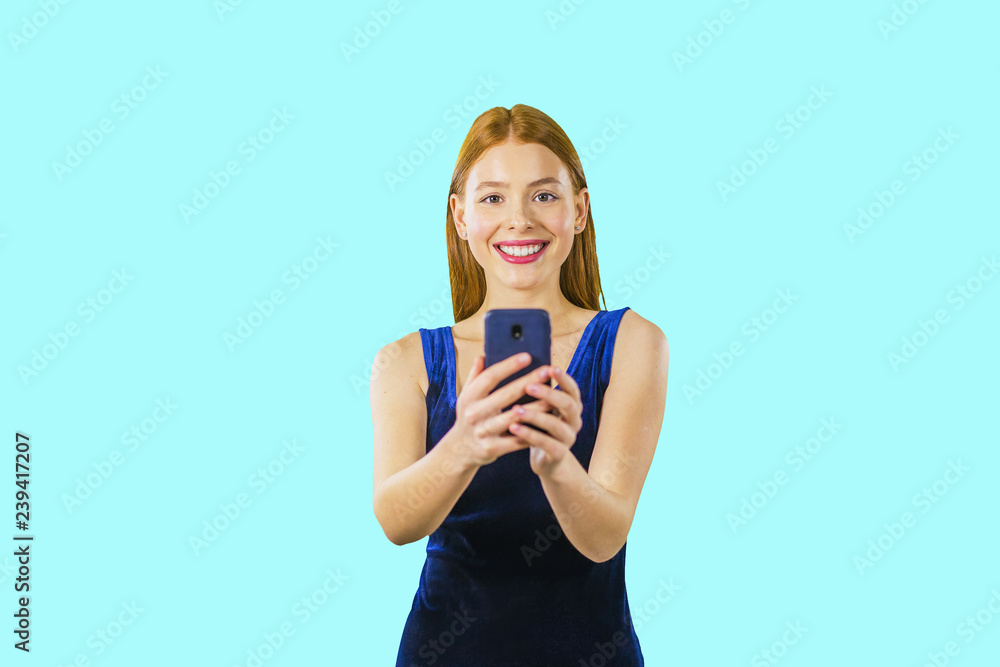 Portrait of a beautiful young red-haired girl in a blue velvet dress with a phone in her hands who is looking at the camera and smiling Portrait of a beautiful young red-haired girl in a blue velvet dress with a phone in her hands who is looking at the camera and smiling