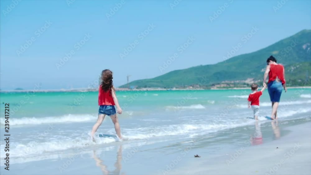 A cute little girl walks along the beach with her mother and little brother, look at the sky on a plane flying by, look at the sky showing hands to the baby.