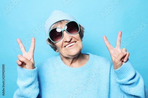 Lifestyle, emotion and people concept: Funny old lady wearing blue sweater, hat and sunglasses showing victory sign. photo