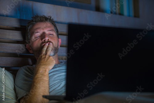 man alone in bed playing cybersex using laptop computer watching porn sex movie late at night with lascivious pervert face expression in internet pornographic sexual content