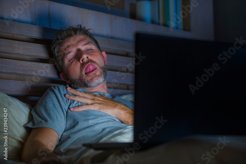 man alone in bed playing cybersex using laptop computer watching porn sex movie late at night with lascivious pervert face expression in internet pornographic sexual content photo