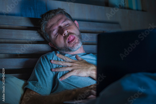 man alone in bed playing cybersex using laptop computer watching porn sex movie late at night with lascivious pervert face expression in internet pornographic sexual content photo