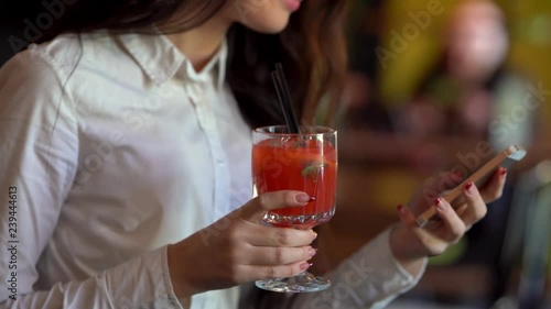 The darkhair girl in white blouse in one hand is holding red cocktail and in the other one her cellphone, looking at cellphone and smiling. photo