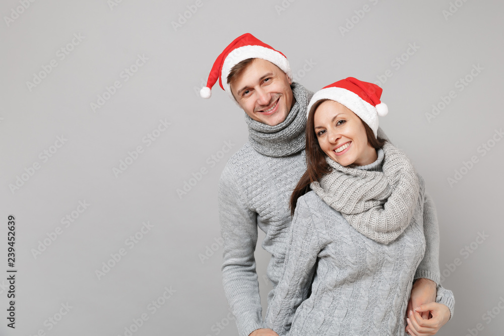 Merry fun couple girl guy in red Santa Christmas hat gray sweaters scarves isolated on grey wall background, studio portrait. Happy New Year 2019 celebration holiday party concept. Mock up copy space. Merry fun couple girl guy in red Santa Christmas hat gray sweaters scarves isolated on grey wall background, studio portrait. Happy New Year 2019 celebration holiday party concept. Mock up copy space.