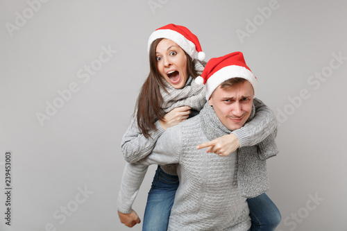 Merry fun couple girl guy in red Santa Christmas hat gray sweaters scarves isolated on grey wall background  studio portrait. Happy New Year 2019 celebration holiday party concept. Mock up copy space.