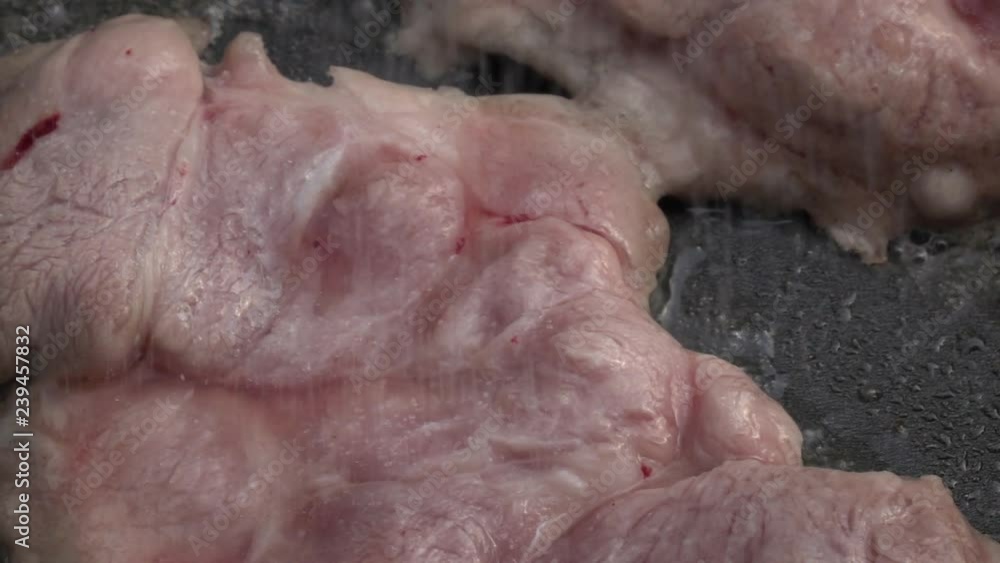 Slow motion of salting sizzling pork steaks on dark gray fry pan with close up. Appetizing natural food background with amazing texture. Mouthwatering home cooking view. High speed camera shooting. Slow motion of salting sizzling pork steaks on dark gray fry pan with close up. Appetizing natural food background with amazing texture. Mouthwatering home cooking view. High speed camera shooting.