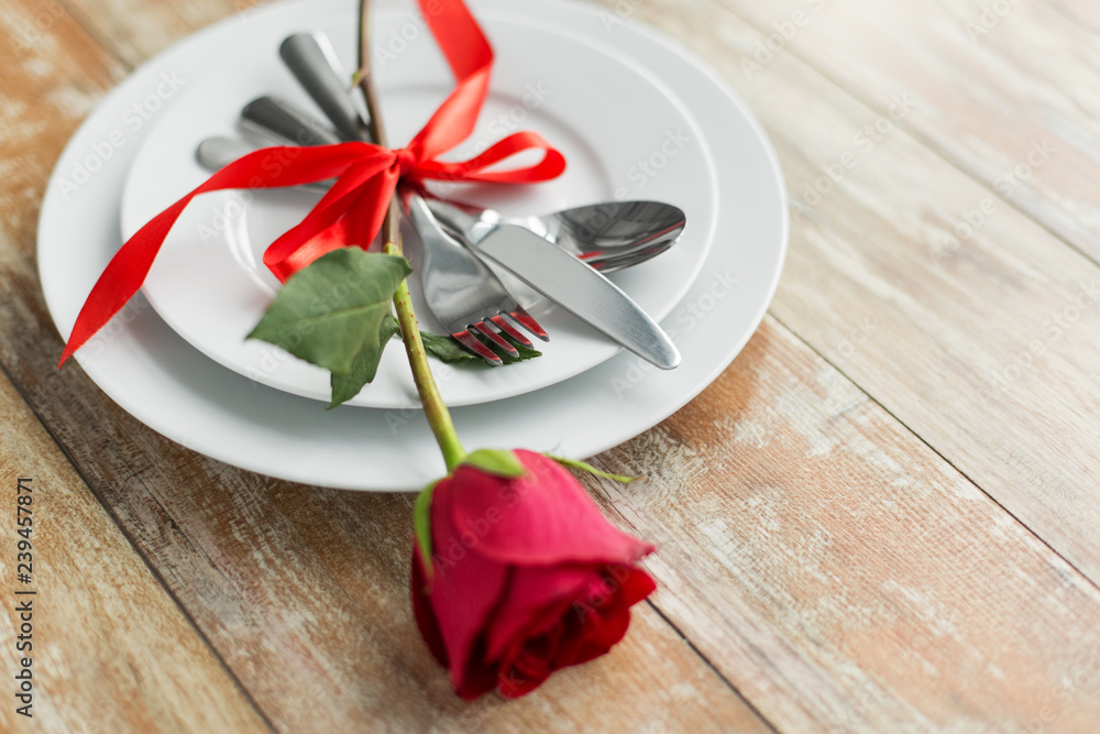 valentines day and romantic dinner concept - close up of red rose flower on set of dishes with cutlery on wooden table valentines day and romantic dinner concept - close up of red rose flower on set of dishes with cutlery on wooden table