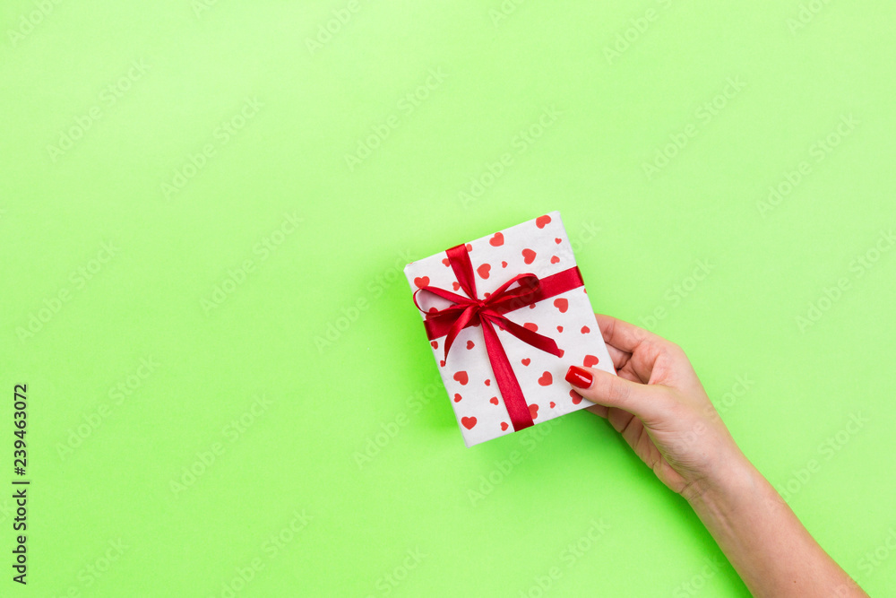 Fototapeta premium Woman hands give wrapped valentine or other holiday handmade present in paper with red ribbon. Present box, red heart decoration of gift on green table, top view with copy space for you design