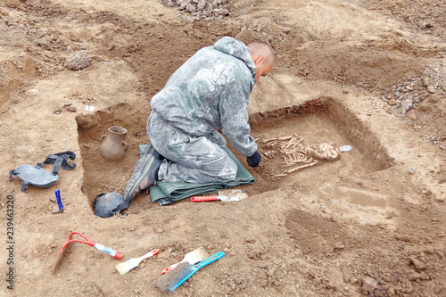 Archaeological excavation. The archaeologist in a digger process, searching tomb, with tools, human bones, part of skeleton and skull in the ground, shovel and brush near. Close up, outdoors.