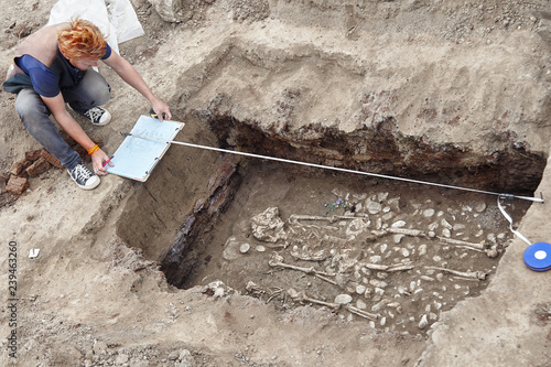 Archaeological excavations. Young stylish archaeologist with red hair makes drowings of human bones, skeleton and skull in the ground tomb. Earphones, mobile phone, sunglass. Outdoors, copy space.