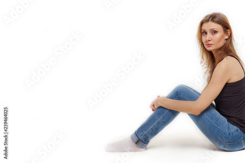 Attractive young caucasian woman with dark hair and eyes sits back to the the wall on the right side of picture, holding her knees with a hands; studio, white background, isolated, copy space.