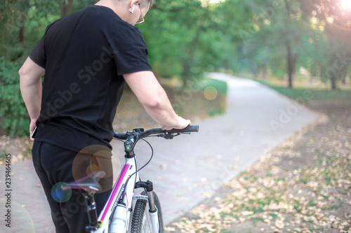 Fototapeta Naklejka Na Ścianę i Meble -  Young caucasian man holds white bicycle in abandoned path in the park, wearing black casual. Earring and earphones; round golden glasses. Outdoors, lenz flair effect. Copy space.