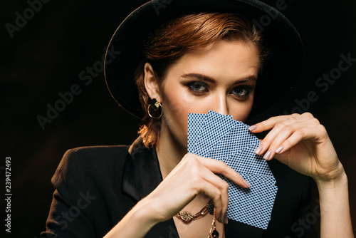 Photos attractive girl in jacket and hat covering face with poker cards isolated on bla