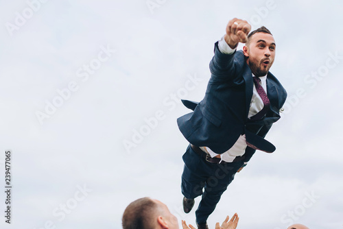 Superman in groom clothes. Flight of the groom. Throw the groom friends. Funny wedding photo. Cheerful bearded groom.