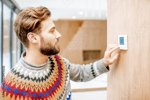 Man in sweater feeling cold adjusting room temperature with electronic thermostat at home photo