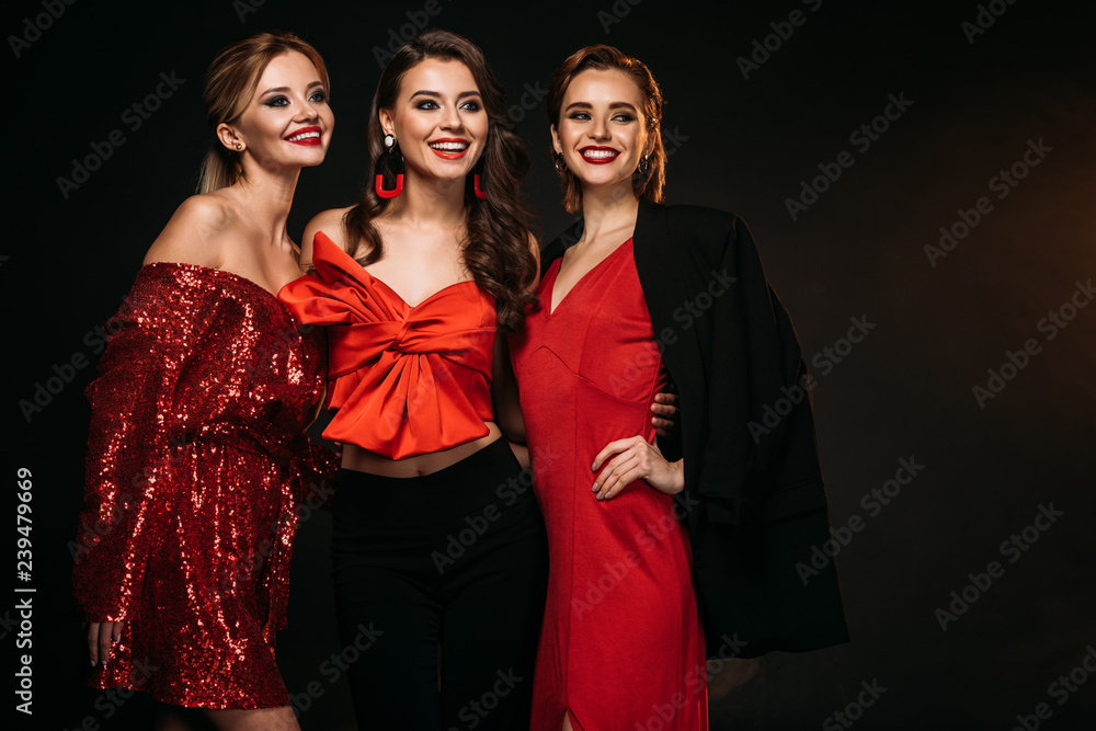 smiling attractive girls in red and black party clothes hugging and looking away isolated on black